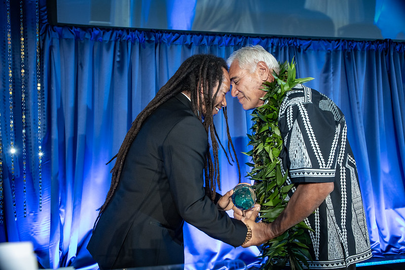 Two men greet each other with clasped hands and foreheads touching as one accepts an award.