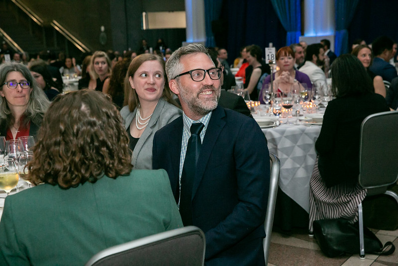 A seated man looks to the side and smiles at a table at an event.